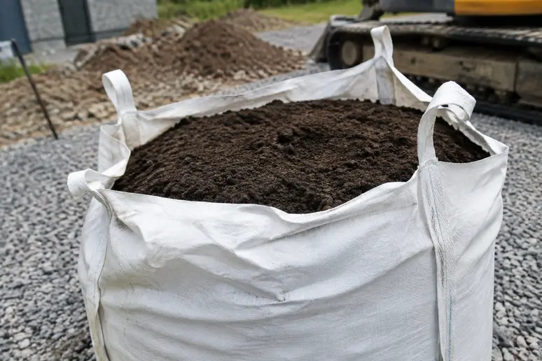 Un big-bag blanc rempli de terre végétale sombre posé sur une cour gravillonnée